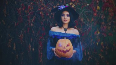 Cadılar Bayramı partisi. Portre fantezisi mutlu gülümseyen kadın cadı. Seksi kız suratı. Klasik mavi kostüm yaratıcı elbise siyah külah şapka ellerinde turuncu balkabağı dumanlı. Sonbahar doğası