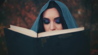 Yakın plan, mistik ortaçağ fantezi kadın cadı okuyucusu. Gotik kız peri kraliçesi büyük bir büyü kitabı okuyor. Akşam makyajı. Mavi, siyah saçları gölgeliyor. Kafasında ipek başlık pelerini var. Sonbahar doğası portakal ağacı
