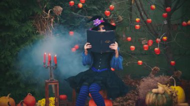 Gizemli fantezi kadın oturur, yüzünü kitabın arkasına saklar. Cadı kız okuyor. Mumlar yanıyor, duman akıyor. Mavi, klasik cadılar bayramı elbisesi, siyah külah şapka. Çıplak ağaç kırmızı elmalar