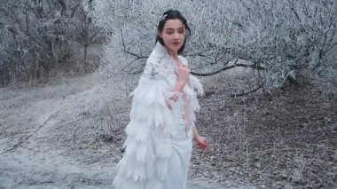 Karnaval kostümü içinde yürüyen güzel bir fantezi kadın Kar Kraliçesi. Beyaz yaratıcı pelerin, gümüş taç, diyabet, kuş tüyü elbise. Yunan tanrıça meleği. Kış doğası, karlı ağaçlar, orman.