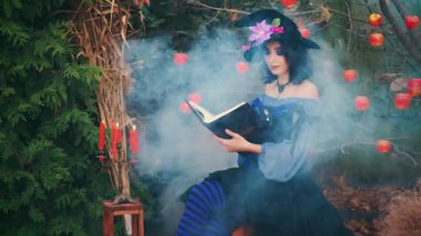 Fantezi kadın elinde kitap tutarak oturur cadı kız büyü okur. Mumlar yanıyor, duman akıyor. Mavi mor karnaval, klasik cadılar bayramı elbisesi, siyah külah şapka. Yeşil ağaç kırmızı elmalar.