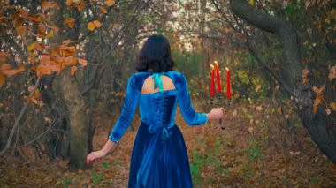 Güzellik sonbahar ormanı, sarı yapraklar, çıplak siyah ağaç gövdeleri. Siluet zarif fantezi kadın. Esmer kadın elinde şamdanla, yanan kırmızı mumlarla sırtını döndü. Uzun mavi eski elbise