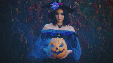 Cadılar Bayramı portresi mutlu gülümseyen kadın cadı. Klasik mor kostümlü, ortaçağ siyah külah şapkalı kız. Elinde turuncu balkabağı tutan gotik kadın. Mavi dumanlı sonbahar balkabağı.