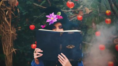 Külahta şapkalı şeytani fantezi kadın yüzünü sihirli kitapların arkasına gizliyor. Büyülü büyüler okuyor. Seksi gözler, yaratıcı mor makyaj. Sonbahar doğası, beyaz duman ağacı kırmızı elmalar. Cadılar Bayramı konsepti