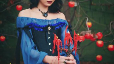  Fantezi kadın cadı elinde antika bir şamdan tutuyor. Yakından mumlar yanıyor, katılaşmış kırmızı balmumu. Sonbahar doğası, orman ağacı, elma meyvesi. Cadılar Bayramı konsepti. Yüzü kesilmiş seksi dudaklar.