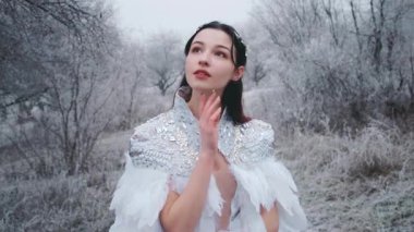 Genç kadın Kar Kraliçesi. Fantezi pelerin, beyaz tüyler. Yaratıcı kıyafetler seksi elbise. Moda modeli, güzel bir yüz. Elf pelerini, kış ormanlarındaki prenses, kışın donan ağaçlar, kar. Gümüş Taç Sirki