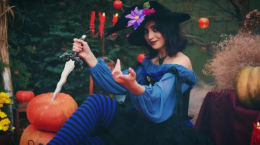 Yaratıcı bıçak tutan güzel bir fantezi kadın. Cadı kız el hareketini davet ediyor. Cadılar Bayramı süslemeleri, balkabakları, elmalar. Mumlar yanıyor, duman akıyor. Mavi klasik elbise, siyah külah şapkalı çiçekler.