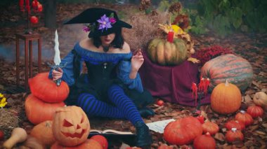 Sanat portresi, cadılar bayramı kadın cadı kostümü elinde eski moda büyü kitabı hançeri ile turuncu balkabağı bayramı dekorunu kesecek. Mavi elbise siyah şapka. Sonbahar doğası kırmızı ağaç bahçesi 