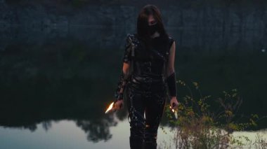 Ellerinde bıçak tutan güzel, fantezi kadın savaşçı. Ateş kılıçları parlak bir şekilde yanar. Kız savunmacı siyah deri ninja yaratıcı kostüm. Yüz maskesi. Manzaralı gece doğa, kayalık nehir kıyısı. 