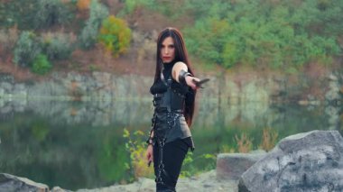 Portre kadını fantezi savaşçısı kamerayı bıçakla tehdit ediyor. Amazon kızı elinde iki hançer tutuyor. Yaratıcı siyah deri seksi kostüm. Uzun saçlı. Sonbahar doğa ağaçları, kayalık nehir kıyısı.