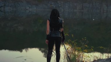 Fantezi kadın savaşçı suikastçı hançeri elinde tutuyor. Gece alacakaranlık manzaralı doğa nehir kayaları. Arkaya bakan seksi kız gizemli yüz maskesi. Siyah deri kostüm, kapüşon. Kılıçlı Ninja. 4k 