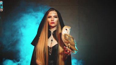 Sanat fantazisi gotik kadın elinde şirin beyaz posta baykuşu kameranın güzel yüzüne bakan dudaklara bakıyor. Masal Kadını Elf Sarışın Seksi Kız. Koyu mavi dumanlı siyah pelerinli açık renk 4k