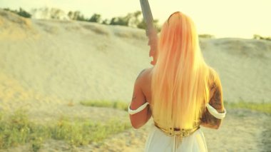 Fantezi kadın kraliçe savaşçı seksi kız güzel güçlü yüz modeli uzun turuncu saçlar arkaya bakıyor. Altın zırh beyaz elbise. Elinde kılıç, kumlu çöl güneşi gökyüzü sanat 4k.