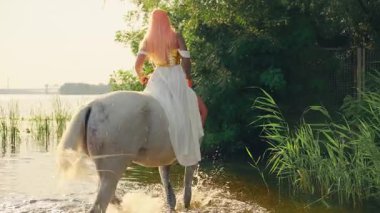 Sanat videosu gerçek insanlar Fantezi kadın savaşçı kraliçe beyaz at toynağına oturur su sıçratır, Yunan tanrıçası nehir gölü boyunca hayvanlara biner, güneş ışığı yeşil gökyüzü ağacı. elf kızı geri dikiz aynası 4k