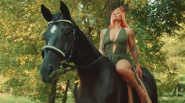 Moda mankeni ata biner. Mutlu fantezi kadın orman perisi at ağızlığını at sırtında okşadı. Peri masalı kızı seksi kızıl saçlı. Yaz ormanı yeşili ağaçlar. 4k