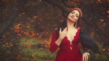 Elinde kocaman siyah bir kuzgun olan güzel esmer kadın portresi. Kırmızı elbiseli kraliçe resmi, zarif saç stili, altın taç ve yakut küpeli. Kamera için poz veriyorum. Sarı yapraklı arka plan ağaçları