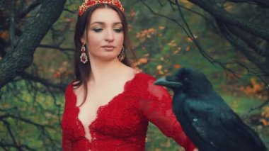 Elinde kocaman siyah bir kuzgun olan güzel esmer kadın portresi. Kırmızı elbiseli kraliçe resmi, zarif saç stili, altın taç ve yakut küpeli. Kamera için poz veriyorum. Sarı yapraklı arka plan ağaçları