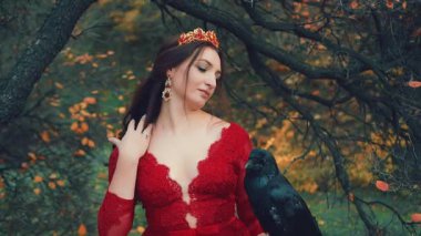 Çekici esmer kadın. Uzun kıvırcık saçlarını altın taç ve yakut küpelerle düzelten kırmızı elbiseli kraliçenin cadılar bayramı görüntüsü. Kuş ötüşüne bakar. Güzel stil sahibi kötü cadı kuzgun.