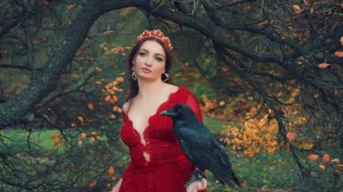 Elinde kocaman siyah bir kuzgun olan güzel esmer kadın portresi. Kırmızı elbiseli kraliçe resmi, zarif saç stili, altın taç ve yakut küpeli. Kamera için poz veriyorum. Sarı yapraklı arka plan ağaçları