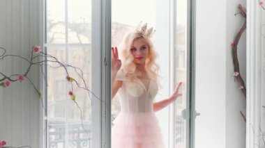 Genç ve güzel prenses şehir manzarasının penceresinde duruyor. Bayan bayan klasik ev odasının içini seviyor. Gizemli gülümseme. Pembe lüks elbise. Uzun sarı kıvırcık saçlı. Eski, parlak taç.