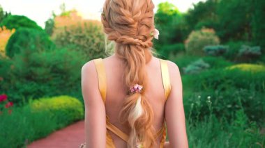 Sarı elbiseli, sırtı açık, uzun örgülü sarışın bir kadın. Yapay saçlı, yaratıcı bir saç modelinin gösterimi. Arka plan kraliyet bahçesi. Yüzsüz, dikiz aynasız çekim