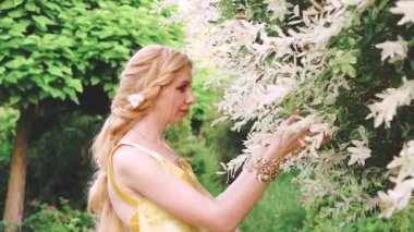 Saçları nazikçe örülmüş çekici, sevimli bir kadın. Çiçek açan bir ağacı okşuyor. Dalları kokluyor ve hafifçe gülümsüyor. Yunan tarzında muhteşem bir orman perisi görüntüsü. Kraliyet bahçesi