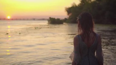 Gri elbiseli, üzgün, yalnız bir kadın gün batımında sırtı kameraya dönük olarak suda duruyor. çok renkli sıcak gökyüzü, güneş nehire yansıyor. Çekici, sevimli peri yürüyüşleri.