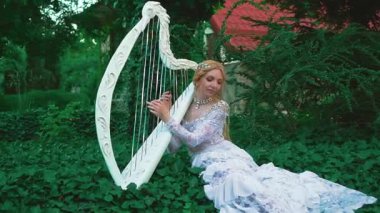 Beyaz, yaratıcı, dantelli, klasik elbiseli çekici sarışın kadın arp çalıyor. Arka plan muhteşem kraliyet bahçesi. Elinde eski bir müzik aleti tutuyor. Kıyafet, kibar bir elf müzisyeninin görüntüsü..
