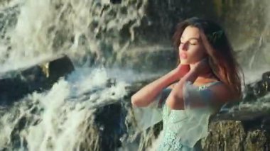 Tatlı, melek gibi bir yüzü olan çekici bir kadın şelalenin arka planında poz veriyor. Rüzgarda dalgalanan saçlar. Hoş bir gülümseme, olumlu duygular. Yaz portresi.