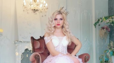 Ortaçağ prensesi yüzünü buruşturur, ağzını ve gözlerini şaşkınlıkla açar. Şirin, güzel kadın yüzü. Eski bir koltukta oturan kız. Beyaz klasik barok odası, çiçekler. Sarışın, kıvırcık saçlı taç