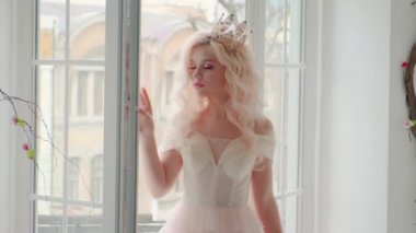 Güzel, genç bir kadın, maket görüntüsü, gizemli prenses. Kafasında lüks bir taç var. Klasik beyaz oda, klasik pencere. Pembe seksi elbise. Uzun kıvırcık dalgalı sarı saçlar. Doğal güzellik.