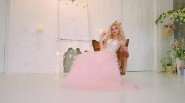 Sandalyede oturan güzel, genç ve çekici bir kadın. Antika ortaçağ pembe gür elbisesi. Klasik oda içi lüks şato. Beyaz stüdyo. Uzun sarı dalgalı dalgalı saçlar altın taç