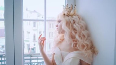 Romantik portre güzel, zarif, üzgün kadın prenses pencerede. Beyaz perde arkası oda içi. Uzun dalgalı dalgalı saçlar. Altın antika taç. Pembe giysili gelin. Peri masalı düğünü 