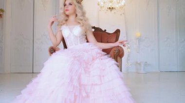 Ortaçağ koltuğunda sıkılmış güzel huysuz prenses. Beyaz oda tarzı barroko. Klasik kristal avize. Sarışın, kıvırcık saçlı kadın. Altın kraliyet tacı. Uzun, lüks, zarif pembe balo elbisesi