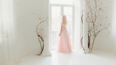 Efsanevi üzgün genç ortaçağ efsunlu prenses klasik cama nazikçe dokunuyor. Uzun, güzel, lüks pembe bir elbise. Sarı dalgalı saçlı bir kadın. Saç stili süslenmiş altın kraliyet tacı. Beyaz oda içi.