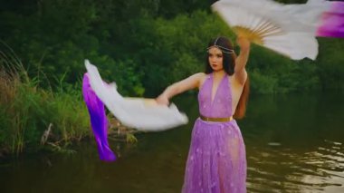 Fantezi perisi el sallıyor. Dans eden iki yelpaze, uzun ipek kumaş rüzgarda uçuşuyor. Yavaş çekimde göbek dansı, mor elbise. Kadın tanrıça elf kraliçesi. Nehir gölü ormanı yeşil ağaçlar yaz doğası 4k