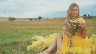 Mutlu kadın yeşil çimlerde oturuyor yaz mevsimi doğa tarlası günbatımı gökyüzü. Sarışın kız elinde bir buket yabani çiçekle alerjiden kurtulmanın tadını çıkarıyor. konsept sağlık dinlenme rahat sarı elbise