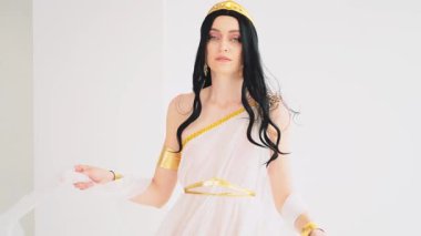 Fantezi Yunan tanrıçası el sallayan, gülen yüzlü kadın. Esmer manken pozu. Seksi kız kraliçe. Beyaz stil ipek kumaş uzun sanat eseri yaratıcı elbise altın taç. yavaş çekim.