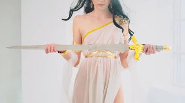 Yunan savaş tanrıçası Athena 'nın antik çelik kılıcı elinde tuttuğu fantezi portresi. Yaratıcı tasarım ipekten elbiseler beyaz yaratıcı elbiseler altın aksesuarlar eski moda kıyafetler rüzgarda uçuşan uzun saçlar