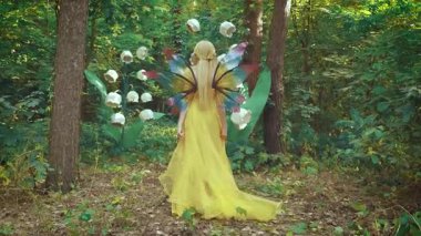 Mutlu fantezi kadın orman perisi. Elf prensesi kız yazın doğa ormanında yürüyor. Sarı elbiseli kelebek kanatları. Büyük çiçekler vadideki beyaz zambak manzarası. Yeşil yapraklar