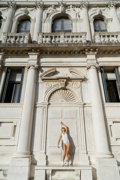 Wide angle view of blonde sportswoman in sunglasses, white sneakers and bodysuit  standing on bench near building in Italy 