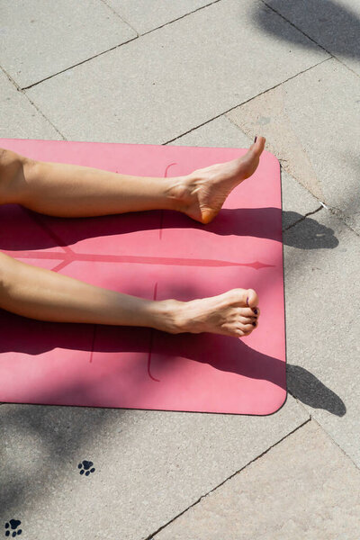 Cropped view of barefoot woman getting tan and lying on yoga mat on urban street, summer concept 