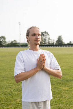 Beyaz pamuklu tişörtlü genç adam Anjali Mudra jesti ile meditasyon yapıyor ve dışarıdaki stadyuma bakıyor.