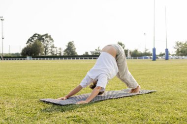 Beyaz tişörtlü, pamuklu pantolonlu, çıplak ayaklı, yunus pozisyonunda yoga yapan, açık havada yeşil çimenlikte duran bir adam.