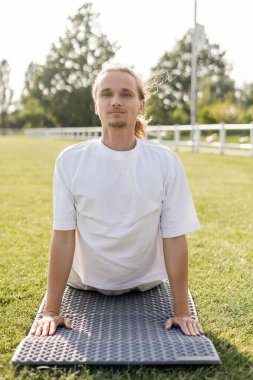 Beyaz tişörtlü genç pozitif adam dışarıda yoga minderinde kobra pozu çalışırken kameraya bakıyor.