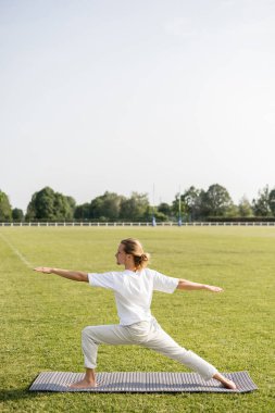 Uzun saçlı ve yalın ayak yoga yapan bir adamın yan görünüşü. Savaşçı pozunda, ellerini çimenli alanda uzatmış.
