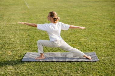 Uzun saçlı, beyaz tişörtlü, savaşçı pozuyla meditasyon yapan, yoga minderinin üzerinde uzanmış genç bir adamın yan görüntüsü.