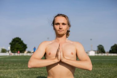 Spor tişörtsüz bir adam kameraya bakıyor ve dışarıda anjali mudra hareketleriyle meditasyon yapıyor.