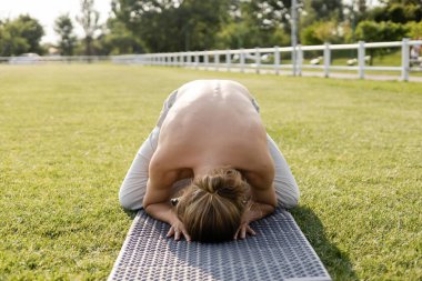 Uzun saçlı üstsüz adam Himalaya ördeği pozunda çimenli tarlada yoga minderinde meditasyon yapıyor.
