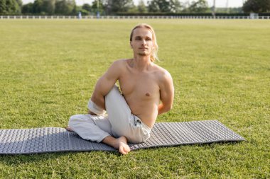 Pamuklu pantolonlu üstsüz adam dışarıda yeşil çimlerde yoga yaparken poz verip gözlerini kaçırıyor.